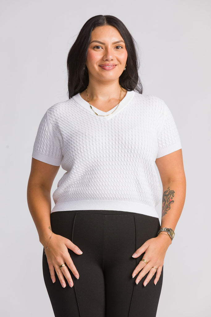 Woman wearing a white textured knit white top and black leggings against a plain background