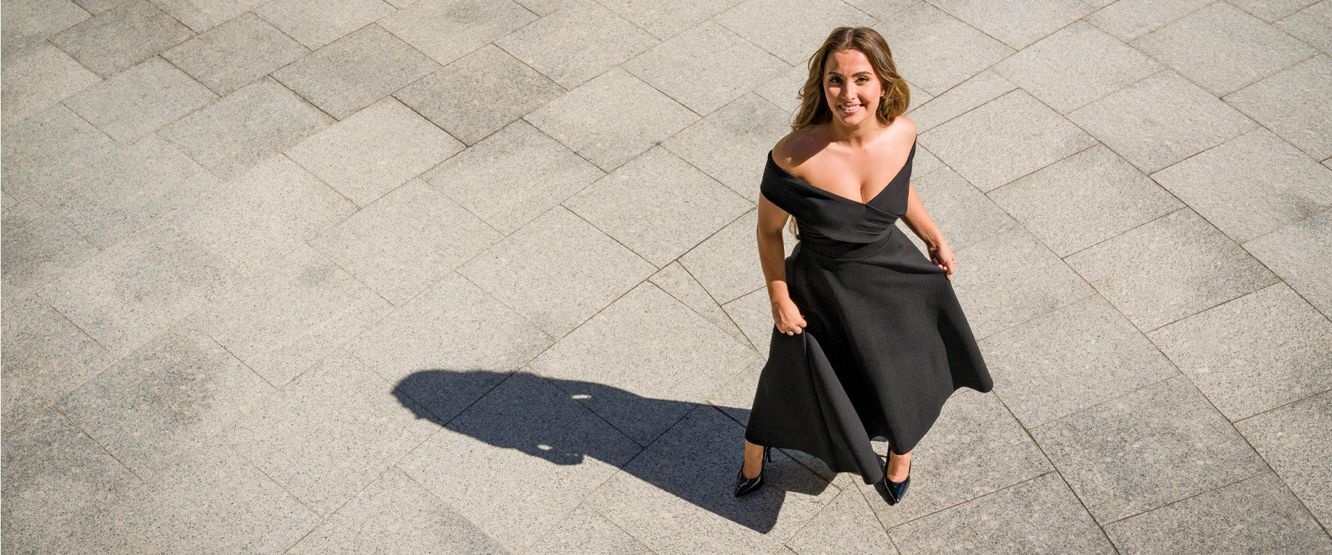 Petite model wearing a black off shoulder midi dress with a fitted bodice and flared skirt, styled in a minimal outdoor setting, designed for petite proportions.