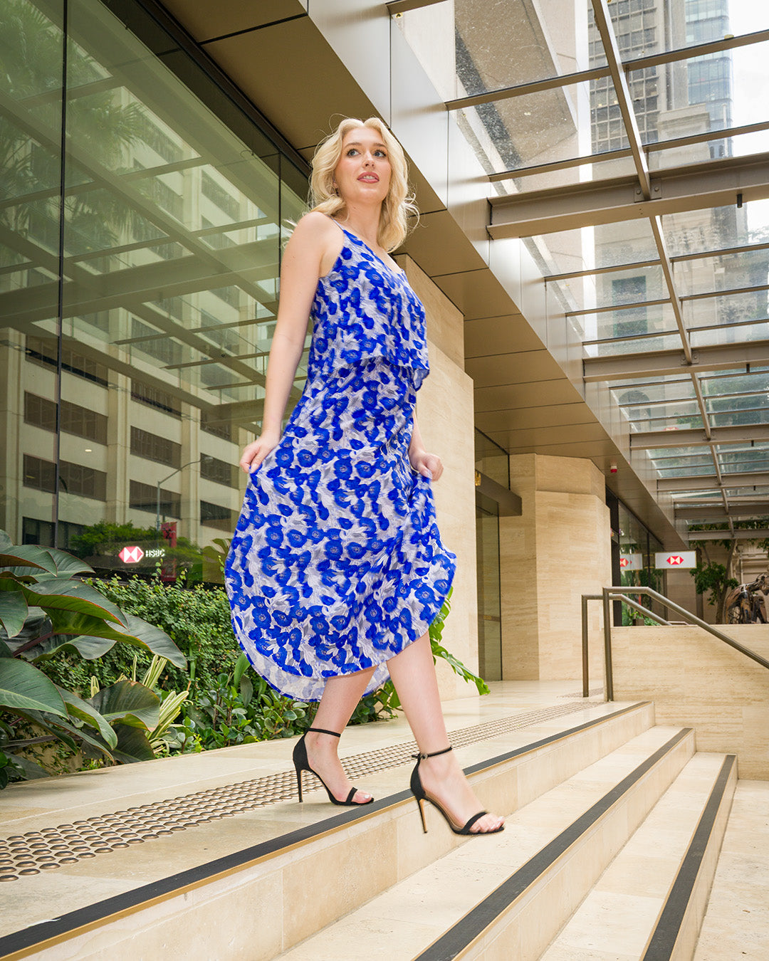 Petite model wearing a blue floral midi dress walking up steps in a modern city setting, highlighting fit and movement for petite proportions.